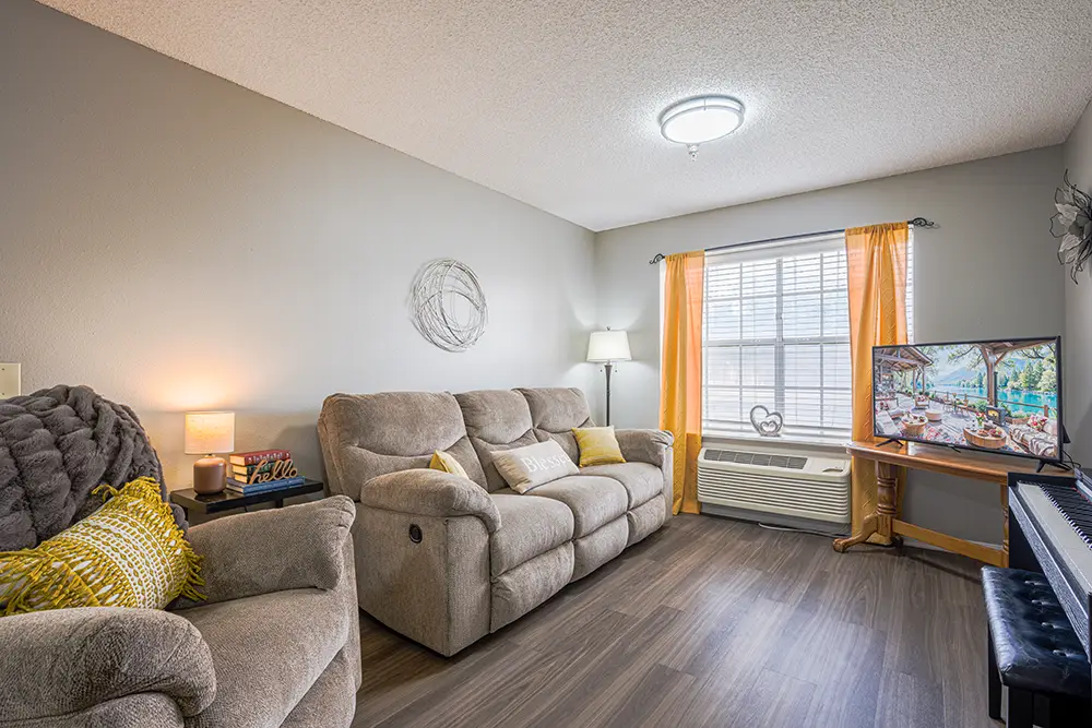 Plum Creek Place-POI-09 A cozy living room with beige reclining sofas, yellow accents, a TV on a stand, a lamp, a window with yellow curtains, and a piano against one wall.