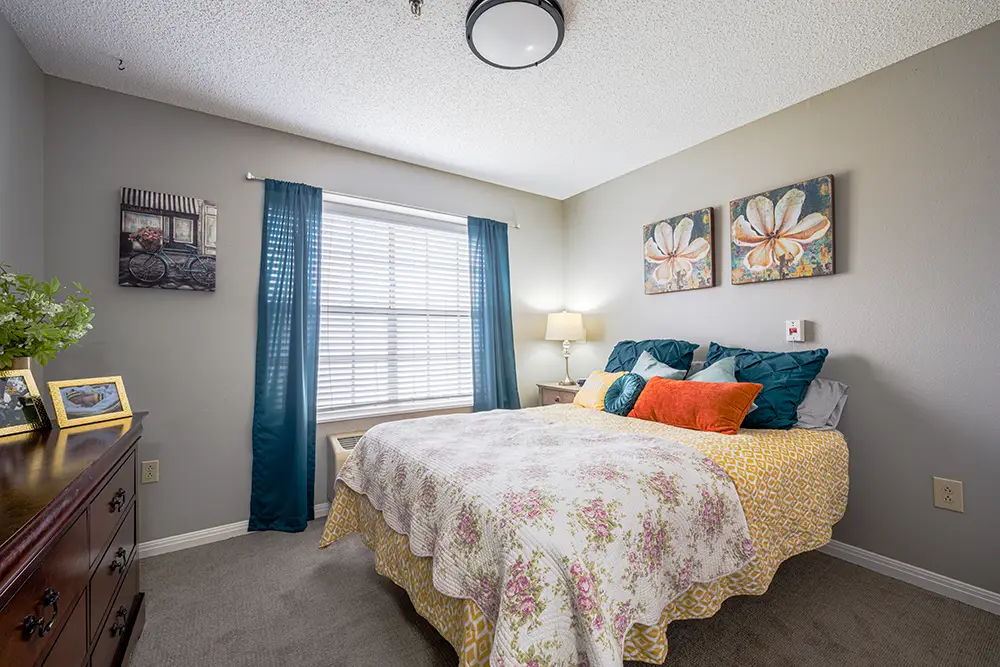 Plum Creek Place-POI-10 A neatly made bed with floral bedding in a bedroom, with blue curtains, wall art, a lamp, and a dresser with framed photos and a plant.