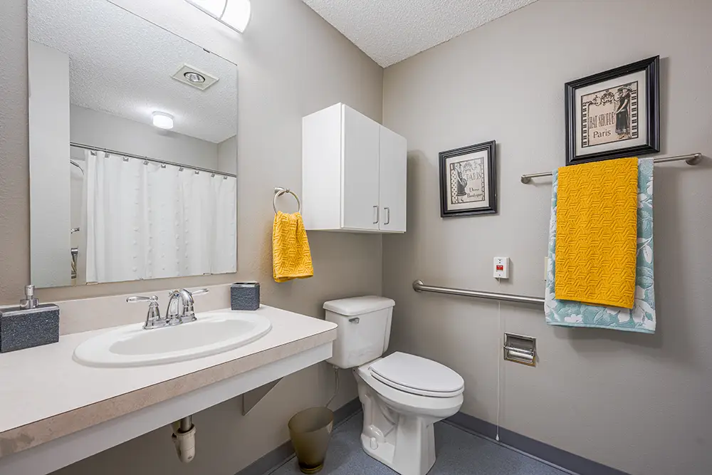Plum Creek Place-POI-11 Accessible bathroom with a wall-mounted sink, toilet, grab bar, white cabinet, towel rack with yellow towel, and framed pictures on a light gray wall.