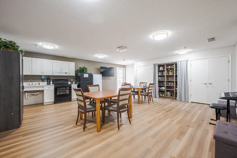Plum Creek Place-POI-12 A spacious room with a kitchenette, dining tables and chairs, bookshelves, and a piano on light wood flooring under ceiling lights.
