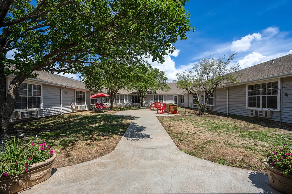 Plum Creek Place-POI-13 A paved walkway leads through a courtyard with grass, trees, red chairs, and a swing, surrounded by one-story white buildings under a blue sky.