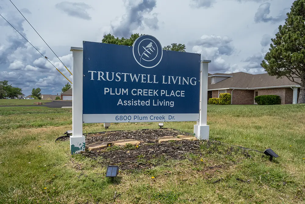 Plum Creek Place-POI-14 Blue and white outdoor sign for Trustwell Living Plum Creek Place Assisted Living, located at 6800 Plum Creek Dr., with buildings and trees in the background.