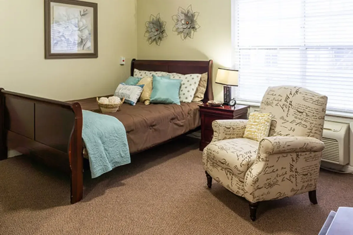A neatly made bed with brown and teal bedding, a large armchair with script print, a side table with a lamp, wall art, and a window with blinds create a warm, welcoming space at Trustwell Senior Living in this carpeted room.