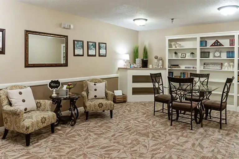 A cozy, carpeted room at Trustwell Senior Living features two armchairs, a round table, a dining table with four chairs, framed art on the walls, and built-in shelves filled with books and decor. A cozy, carpeted room at Trustwell Senior Living features two armchairs, a round table, a dining table with four chairs, framed art on the walls, and built-in shelves filled with books and decor.