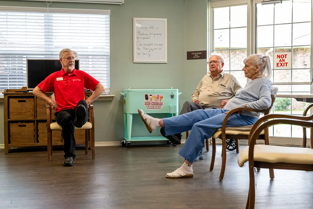 Three older adults sit in chairs, lifting one leg each, participating in a seated exercise class in a well-lit room with windows and a whiteboard. Three older adults sit in chairs, lifting one leg each, participating in a seated exercise class in a well-lit room with windows and a whiteboard.