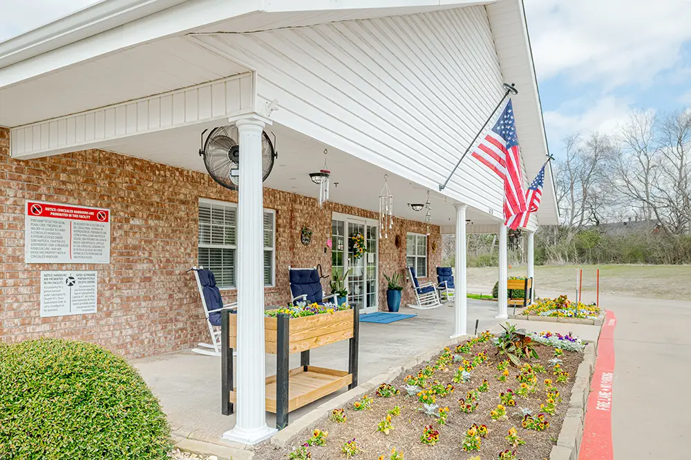 Brick building with white columns, American flags, rocking chairs, a flower bed, and posted signs outside the entrance on a cloudy day.