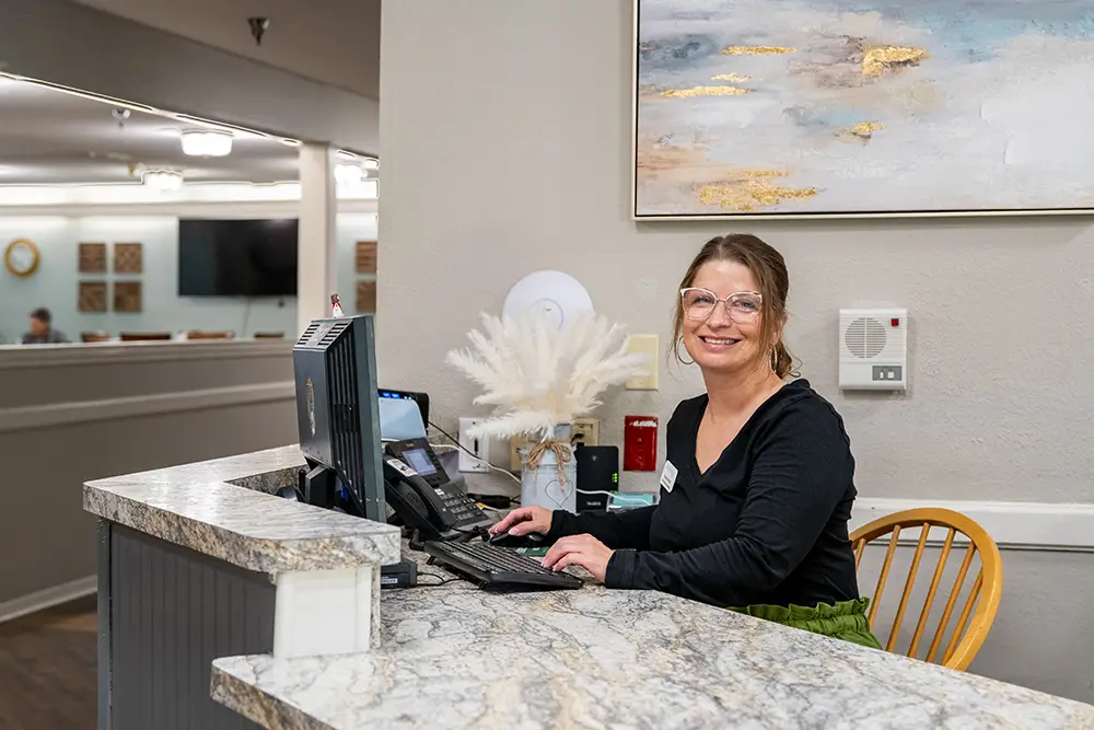 A woman wearing glasses sits at a reception desk with a computer, smiling at the camera in a well-lit office or lobby area. A woman wearing glasses sits at a reception desk with a computer, smiling at the camera in a well-lit office or lobby area.