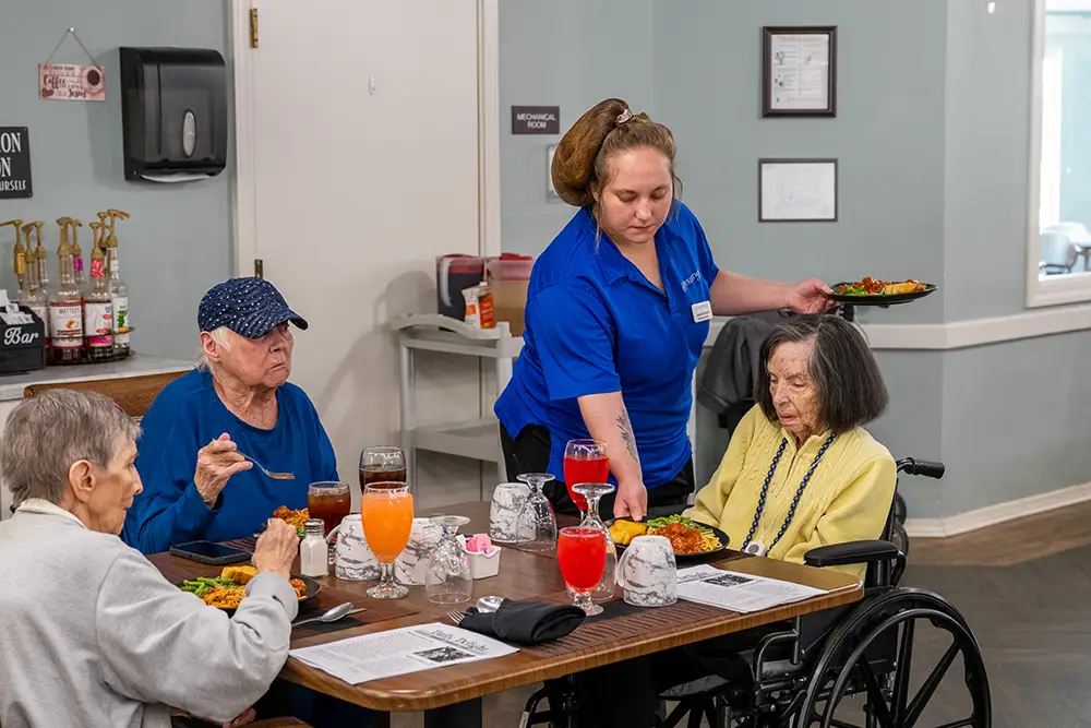 A staff member in blue serves food to three elderly women, one in a wheelchair, seated at a table with meals and drinks in a care facility dining area. A staff member in blue serves food to three elderly women, one in a wheelchair, seated at a table with meals and drinks in a care facility dining area.