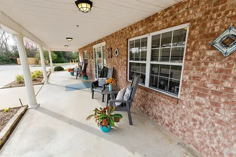 A covered brick front porch at Trustwell Senior Living features rocking chairs, potted plants, and decorative wall hangings beside large windows. A covered brick front porch at Trustwell Senior Living features rocking chairs, potted plants, and decorative wall hangings beside large windows.