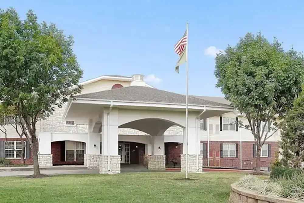 A two-story brick and stone Trustwell Senior Living building with a covered entrance, two flags on poles, and trees on either side, set against a blue sky.