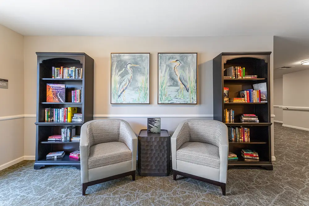 Two armchairs sit between bookshelves and a small table, with two bird paintings hanging on the wall above. Two armchairs sit between bookshelves and a small table, with two bird paintings hanging on the wall above.