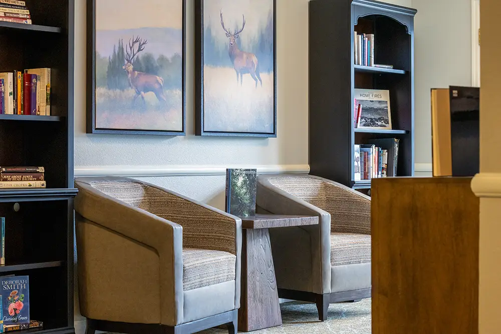 Two gray armchairs and a small table sit between bookshelves, with two large deer paintings on the wall behind them. Two gray armchairs and a small table sit between bookshelves, with two large deer paintings on the wall behind them.