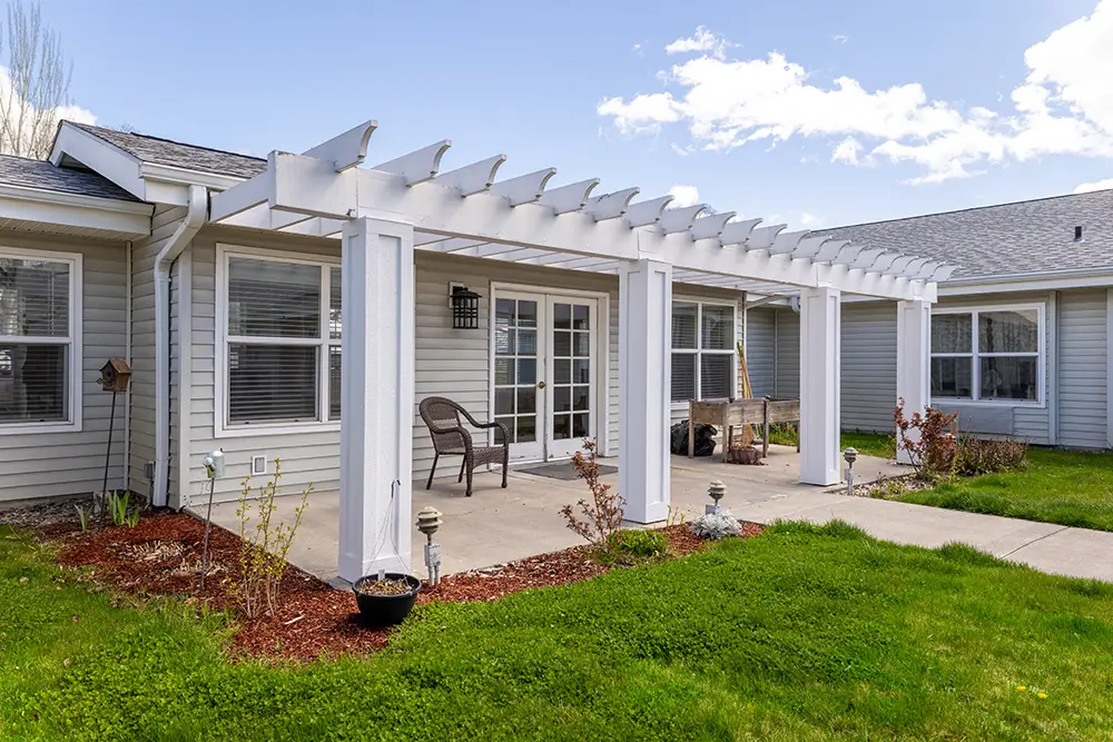 A backyard patio features a white pergola, two chairs, a bench, and a small table outside double glass doors. The house has light siding and trimmed lawn with small shrubs. A backyard patio features a white pergola, two chairs, a bench, and a small table outside double glass doors. The house has light siding and trimmed lawn with small shrubs.