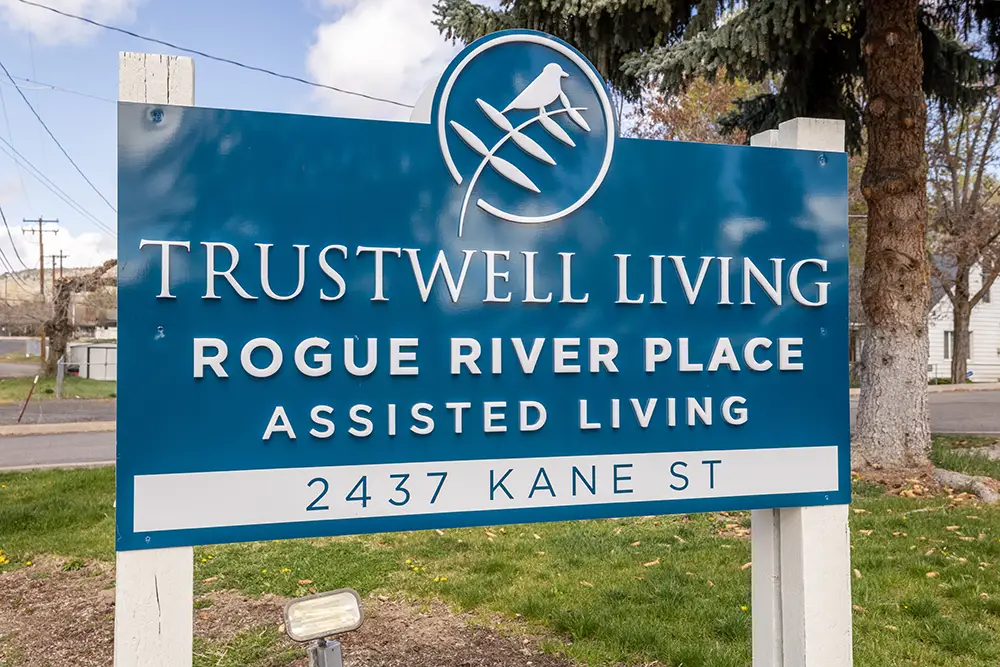 Blue and white sign for Trustwell Living Rogue River Place Assisted Living, located at 2437 Kane St, stands outdoors near a tree. Blue and white sign for Trustwell Living Rogue River Place Assisted Living, located at 2437 Kane St, stands outdoors near a tree.