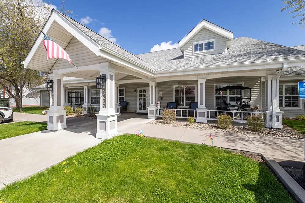 Single-story building with a covered entrance, American flag, porch chairs, and a well-maintained lawn under a clear blue sky. Single-story building with a covered entrance, American flag, porch chairs, and a well-maintained lawn under a clear blue sky.