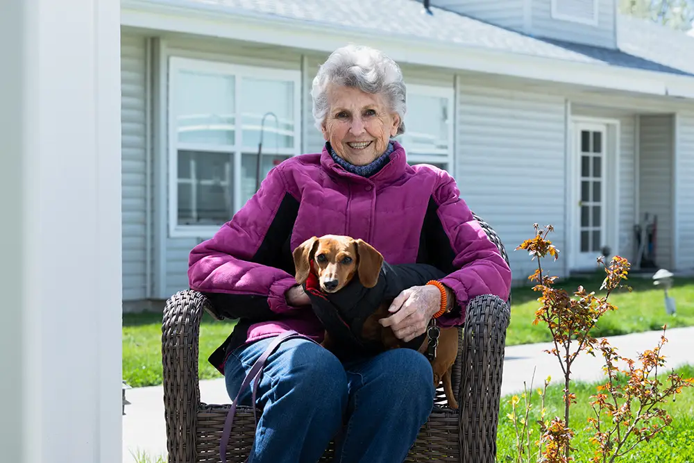 An elderly woman wearing a purple jacket sits on a wicker chair outside, smiling and holding a small brown dog on her lap. A house and a plant are visible in the background. An elderly woman wearing a purple jacket sits on a wicker chair outside, smiling and holding a small brown dog on her lap. A house and a plant are visible in the background.