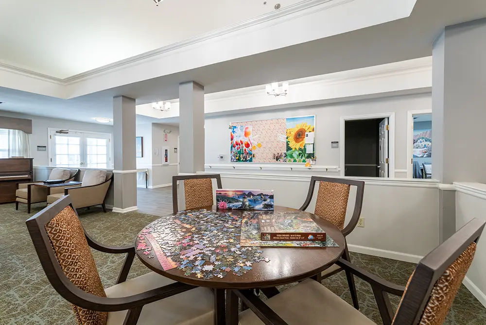 A round table with a jigsaw puzzle in progress is surrounded by four chairs in a bright, carpeted room with armchairs, large windows, and colorful artwork on the wall. A round table with a jigsaw puzzle in progress is surrounded by four chairs in a bright, carpeted room with armchairs, large windows, and colorful artwork on the wall.