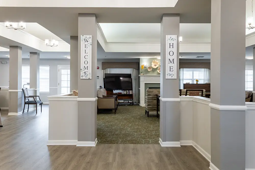 Spacious living area with a TV, armchairs, and fireplace; signs on pillars read “Welcome” and “Home”; light grey walls and wood flooring. Spacious living area with a TV, armchairs, and fireplace; signs on pillars read "Welcome" and "Home"; light grey walls and wood flooring.