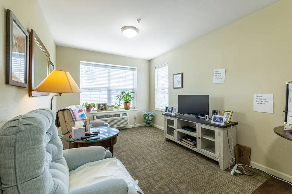 A small living room with beige walls, a TV on a stand, armchairs, framed pictures, a window with blinds, an air conditioner, a lamp, and a potted plant. A small living room with beige walls, a TV on a stand, armchairs, framed pictures, a window with blinds, an air conditioner, a lamp, and a potted plant.