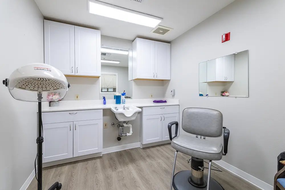 A small salon room with a gray styling chair, hooded hair dryer, white cabinets, a sink, countertop items, and wall mirrors. A small salon room with a gray styling chair, hooded hair dryer, white cabinets, a sink, countertop items, and wall mirrors.