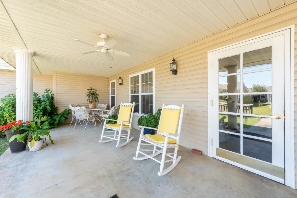 A covered patio at Trustwell Senior Living features two yellow rocking chairs, a glass door, large windows, potted plants, and a small dining table set in the corner.