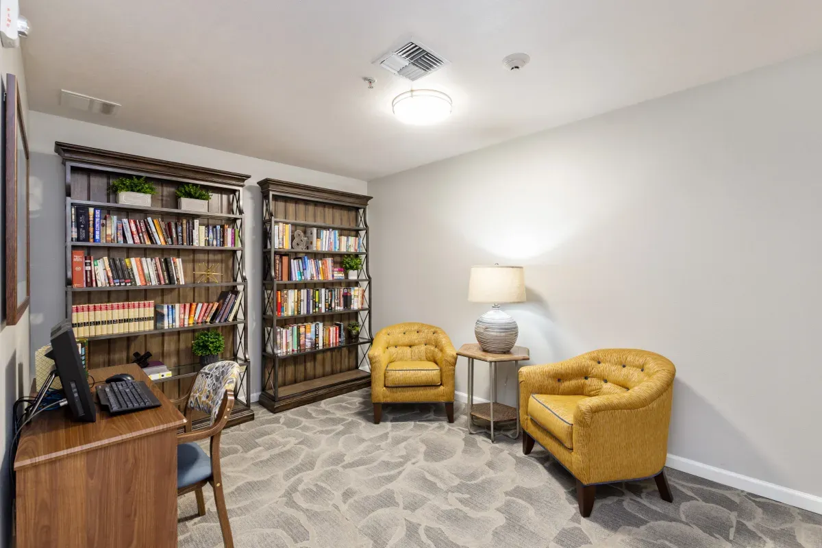 A cozy library room at Trustwell Senior Living features two yellow armchairs, a round side table with a lamp, bookshelves filled with books, and a desk with a computer and chair—perfect for relaxation and quiet reading.