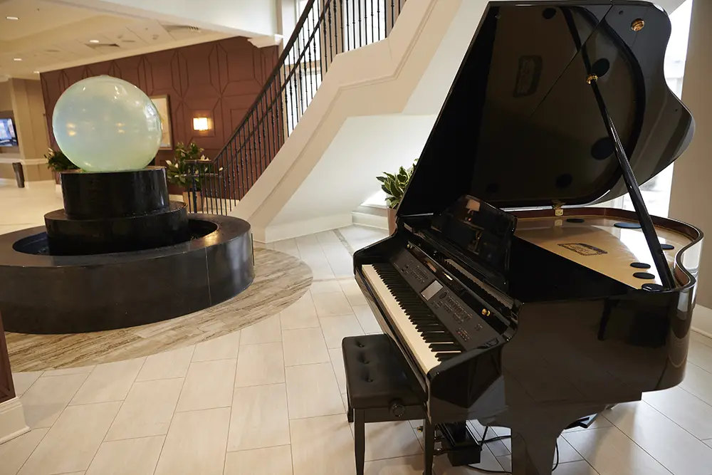 TWRH1 A black grand piano with a bench is placed near a black water fountain with a large glowing orb in a hotel lobby with tiled floors and a staircase.