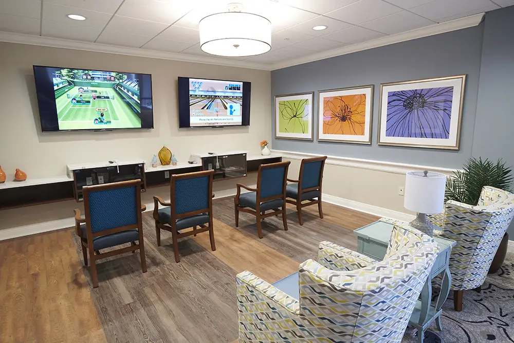 TWRH11 A room with six chairs facing two large wall-mounted TVs displaying video games, colorful flower artwork on the wall, and a modern ceiling light.