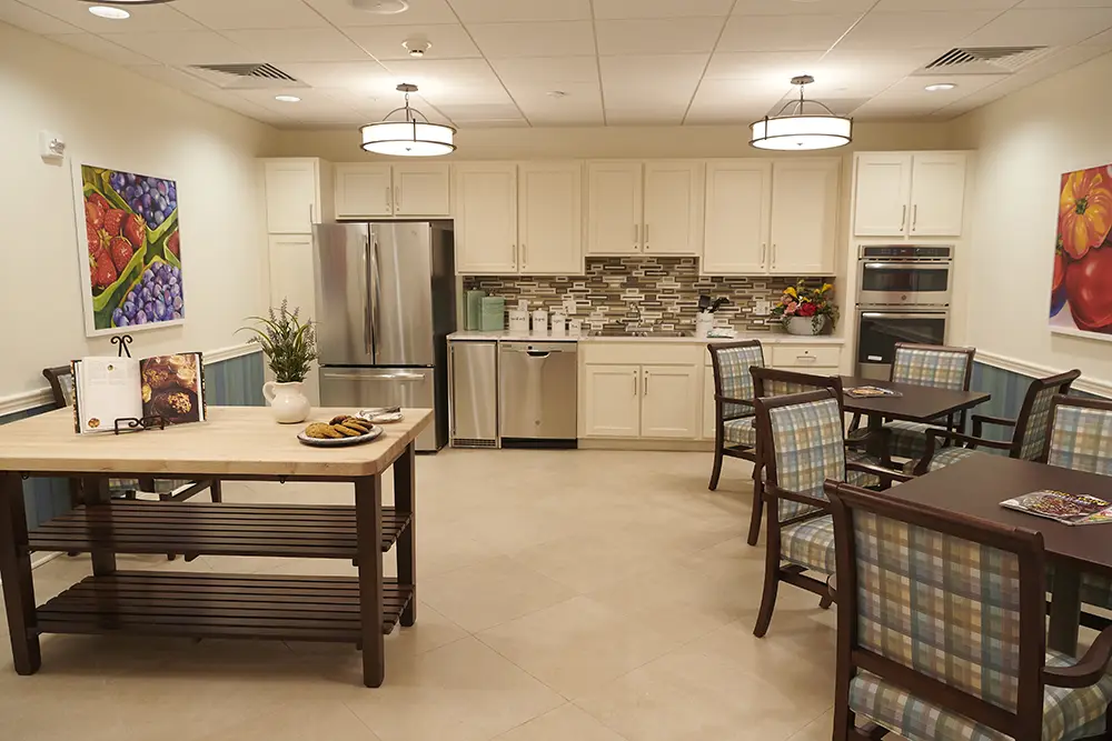 TWRH12 A modern kitchen with white cabinets, stainless steel appliances, a wooden island with cookies, two tables with plaid chairs, and colorful fruit artwork on the walls.