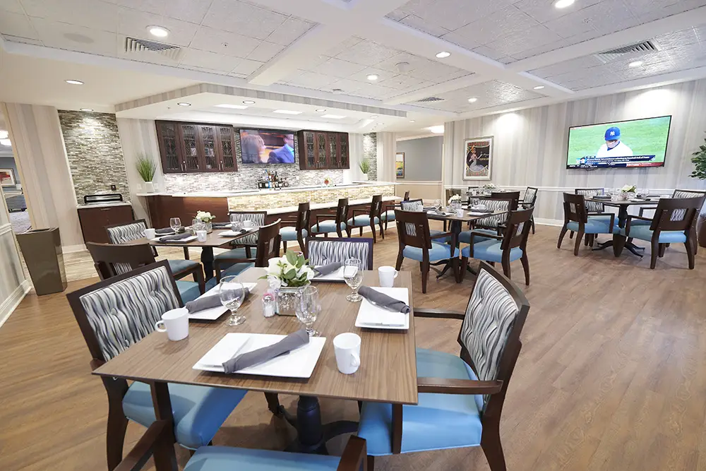 TWRH13 A modern dining room with set tables, wooden chairs with blue cushions, a bar area, and a TV on the wall displaying a baseball game.