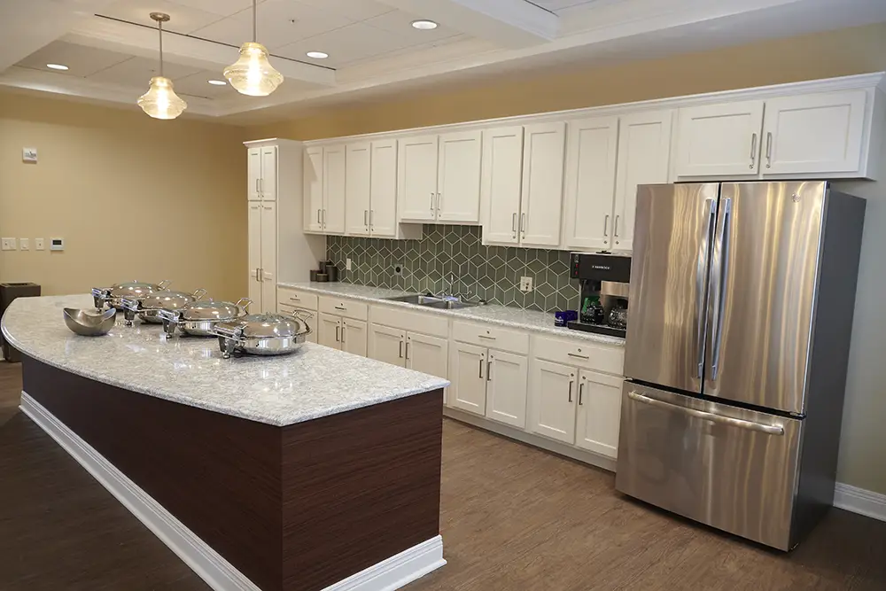 TWRH15 Modern kitchen with white cabinets, stainless steel French door fridge, marble countertops, and a curved buffet counter with chafing dishes. Green tile backsplash and wooden floor.