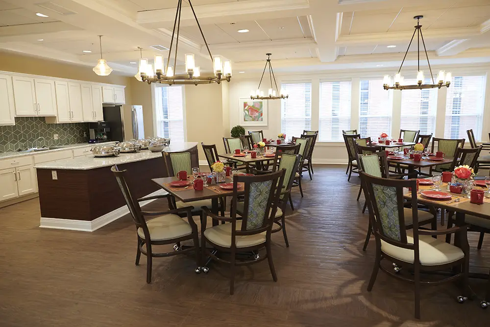 TWRH16 A well-lit dining area with round tables and chairs, set with plates and glasses; a buffet counter and kitchen space are visible in the background.