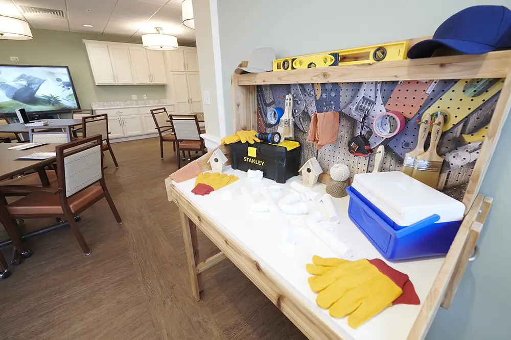 TWRH17 A wooden workbench with gloves, tools, birdhouses, and supplies is set up in a room with tables, chairs, and a TV in the background.