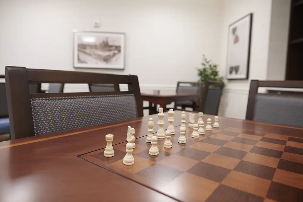 TWRH3 A chessboard with white chess pieces set up for a game sits on a wooden table in a quiet, well-lit room with chairs and framed artwork on the walls.