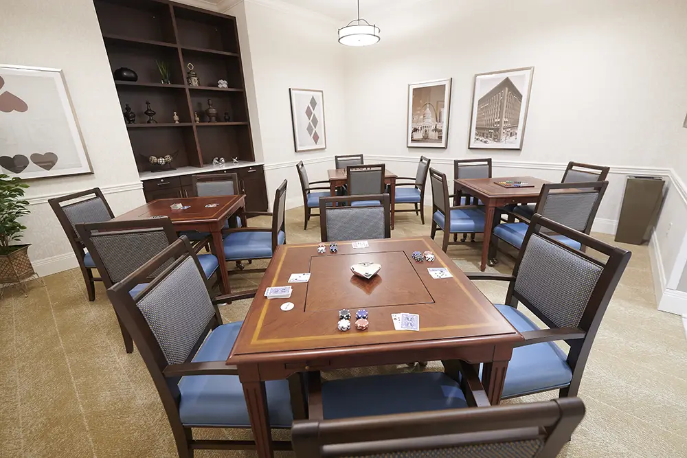 TWRH4 A well-lit room with four wooden tables, each set with poker chips and playing cards, surrounded by blue-cushioned chairs and decorated with framed art and shelving.