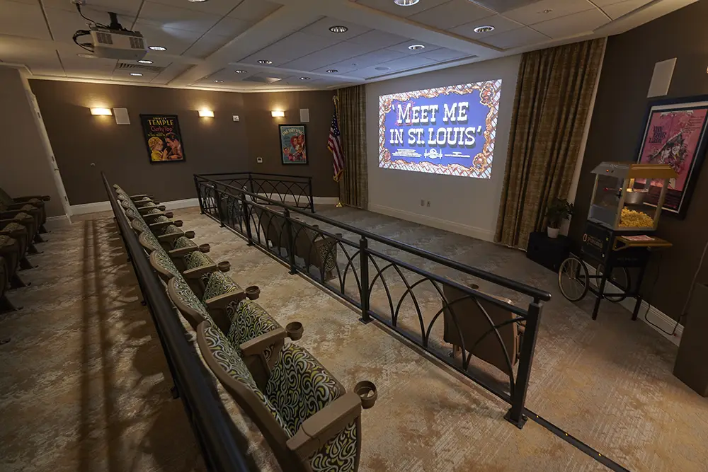 TWRH5 Small private theater room with patterned chairs, a projector screen displaying “Meet Me in St. Louis,” an American flag, and a popcorn machine on the right side.