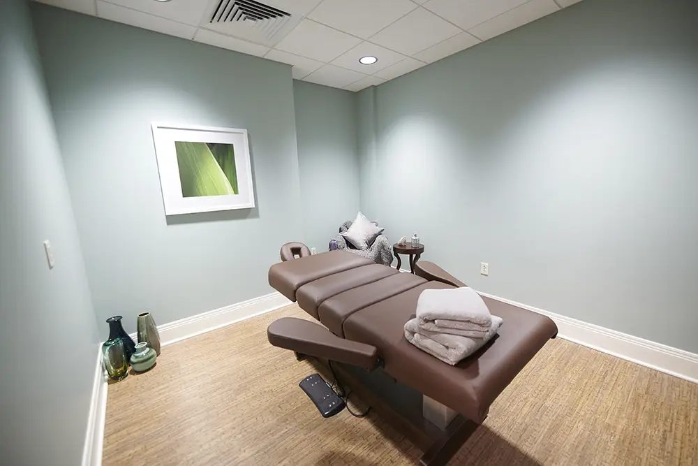 TWRH7 A massage room with a brown massage table, folded towels, a chair with pillows, decorative vases, and a framed leaf artwork on a light blue wall.