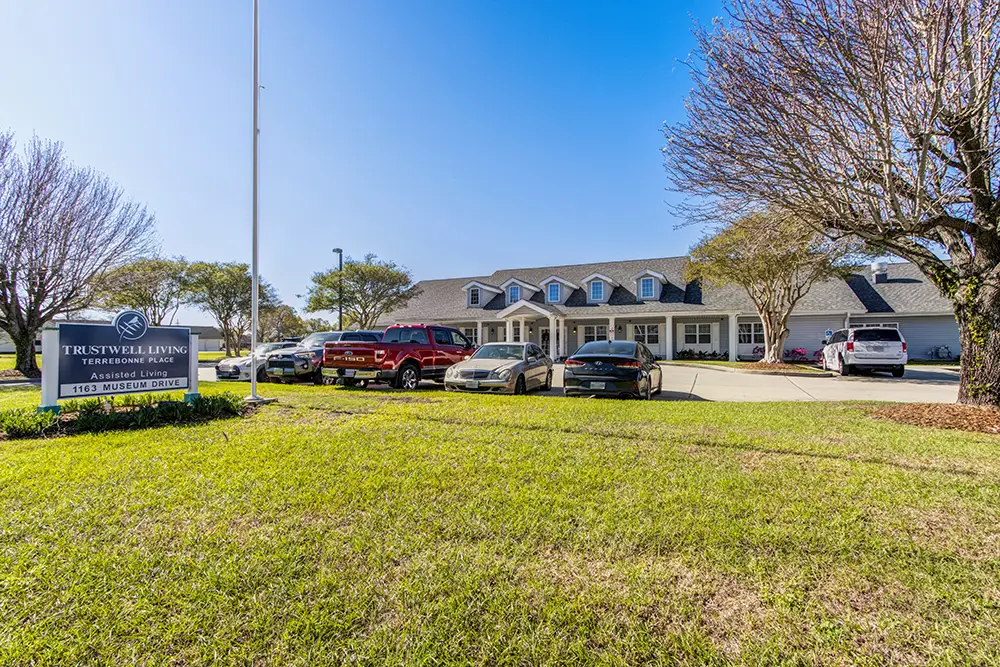 A single-story assisted living facility with multiple cars parked in front, trees lining the driveway, and a sign reading “Trustwell Living Residence Place” on a sunny day. A single-story assisted living facility with multiple cars parked in front, trees lining the driveway, and a sign reading "Trustwell Living Residence Place" on a sunny day.