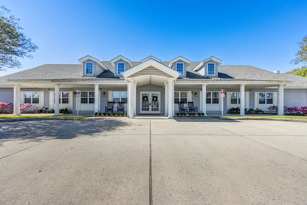Single-story building with a covered entrance, white columns, gray roof, dormer windows, and rocking chairs on the porch, set against a clear blue sky. Single-story building with a covered entrance, white columns, gray roof, dormer windows, and rocking chairs on the porch, set against a clear blue sky.