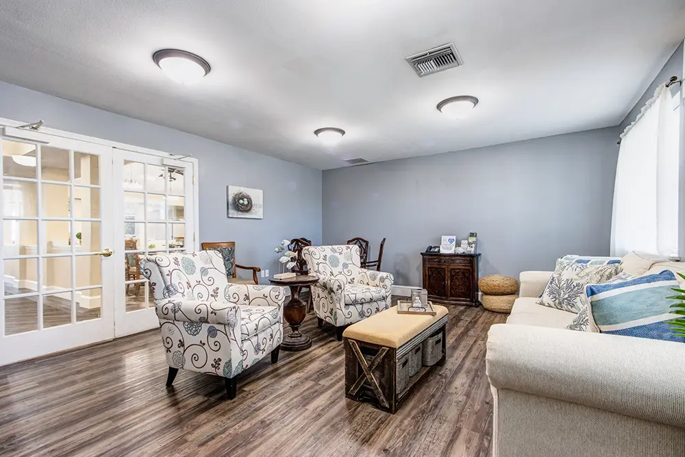 Living room with wood flooring, patterned armchairs, cream sofa, glass double doors, and a bench-style coffee table; neutral color palette with blue accents and natural light from windows. Living room with wood flooring, patterned armchairs, cream sofa, glass double doors, and a bench-style coffee table; neutral color palette with blue accents and natural light from windows.