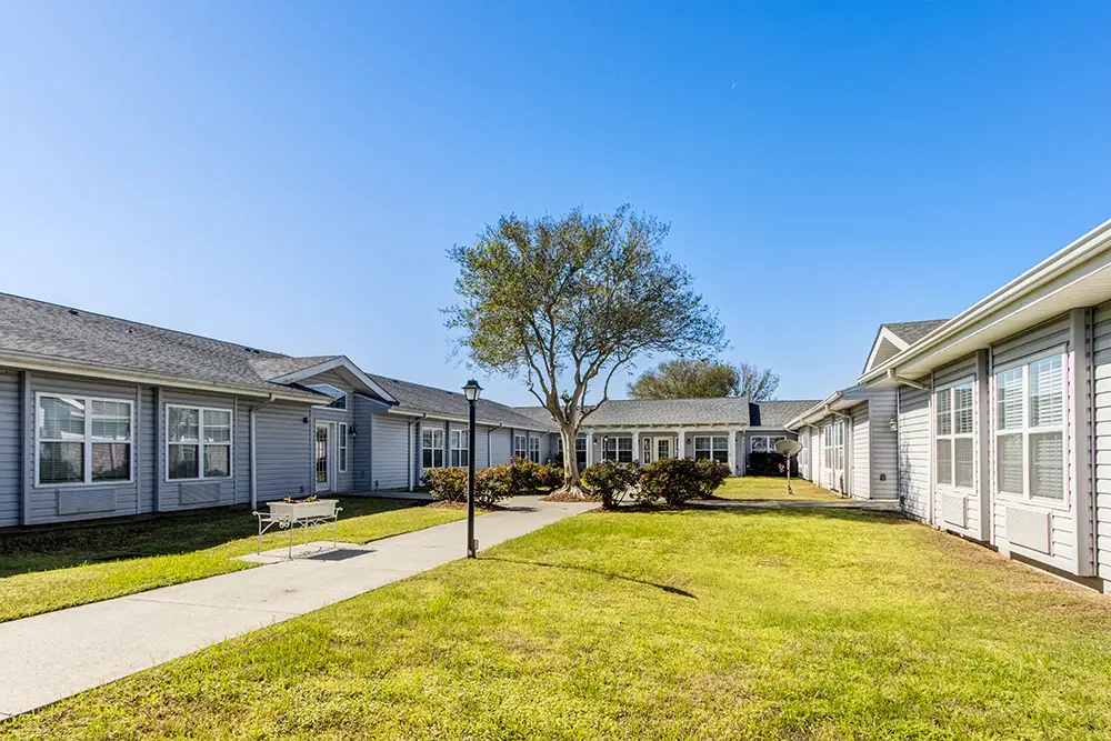 A path runs through a grassy courtyard surrounded by single-story gray buildings with many windows under a clear blue sky. A path runs through a grassy courtyard surrounded by single-story gray buildings with many windows under a clear blue sky.