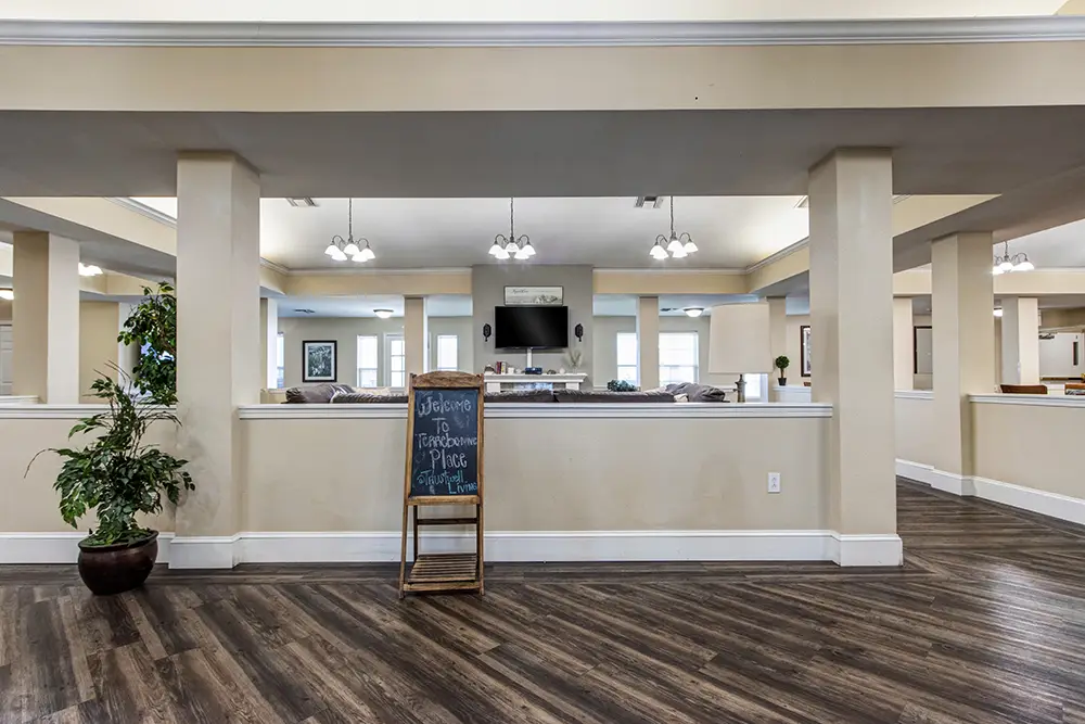 A large open room with beige pillars, wood flooring, a chalkboard sign, indoor plants, and ceiling lights. A TV is mounted on the far wall. A large open room with beige pillars, wood flooring, a chalkboard sign, indoor plants, and ceiling lights. A TV is mounted on the far wall.