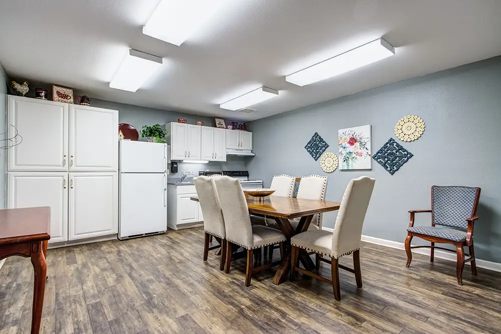A kitchen with white cabinets, a refrigerator, stove, and a wooden dining table with six upholstered chairs. Wall art and a single armchair decorate the gray and blue walls. A kitchen with white cabinets, a refrigerator, stove, and a wooden dining table with six upholstered chairs. Wall art and a single armchair decorate the gray and blue walls.