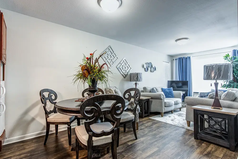 Modern living and dining area with elegant chairs, round table, sofas, lamps, wall art, wood flooring, and natural light from large window with blue curtains. Modern living and dining area with elegant chairs, round table, sofas, lamps, wall art, wood flooring, and natural light from large window with blue curtains.