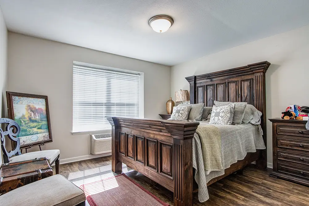 A bedroom with a large wooden bed, several pillows, a nightstand with decor, a framed painting against the wall, and sunlight streaming through a window with blinds. A bedroom with a large wooden bed, several pillows, a nightstand with decor, a framed painting against the wall, and sunlight streaming through a window with blinds.