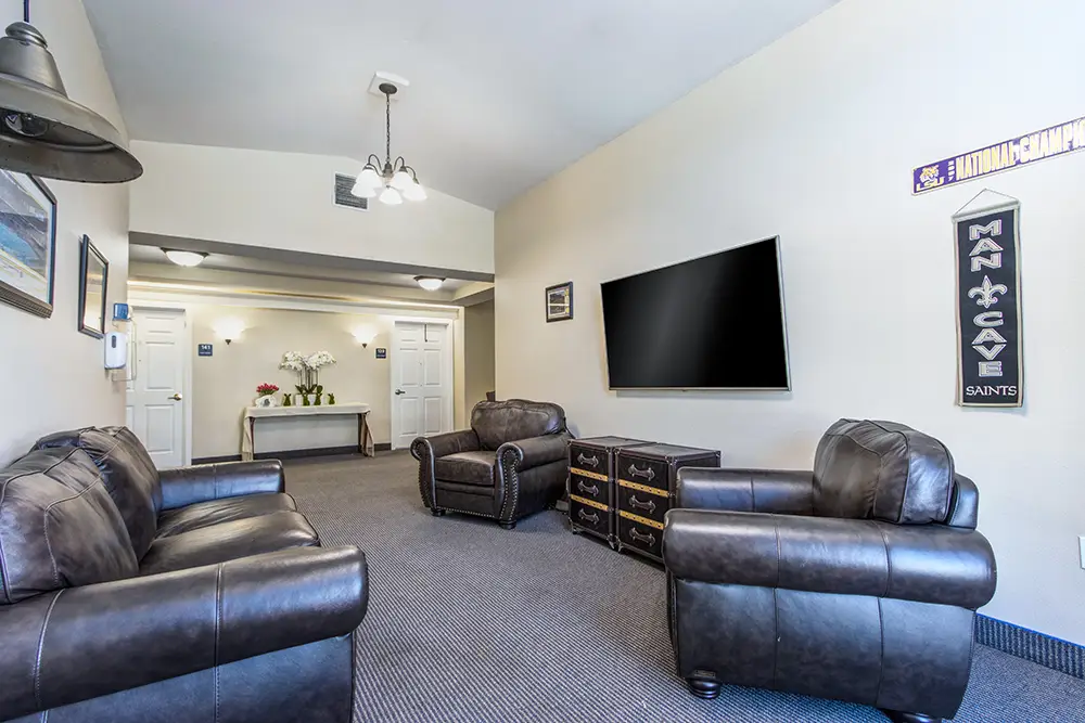 Room with leather sofas and armchairs, a large wall-mounted TV, sports-themed decor, gray carpet, and white double doors in the background. Room with leather sofas and armchairs, a large wall-mounted TV, sports-themed decor, gray carpet, and white double doors in the background.