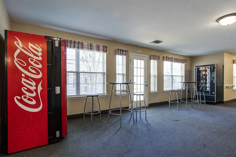 Room with a Coca-Cola vending machine, a snack vending machine, tall round tables with metal stools, large windows, and carpeted floor. Room with a Coca-Cola vending machine, a snack vending machine, tall round tables with metal stools, large windows, and carpeted floor.