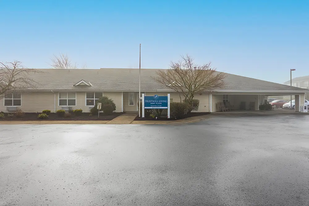 Single-story building with a gray roof, leafless trees, and a blue Trustwell Living sign in front at Kent Place, viewed from a wet, empty parking lot under a clear blue sky. Single-story building with a gray roof, leafless trees, and a blue Trustwell Living sign in front at Kent Place, viewed from a wet, empty parking lot under a clear blue sky.