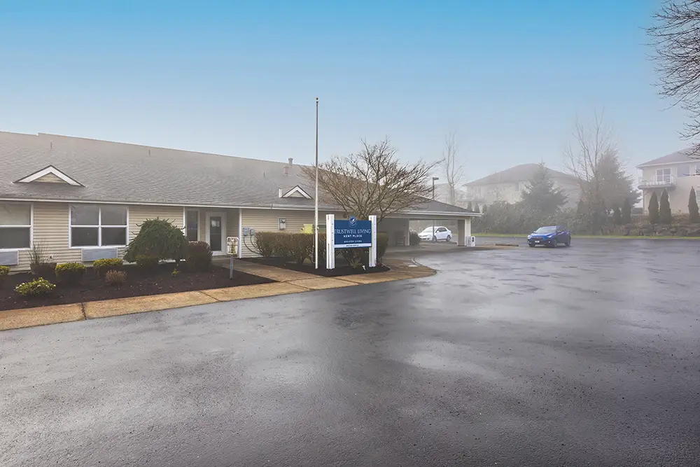 A single-story building with a sign in front, surrounded by a parking lot with one blue car, under a foggy sky—a welcoming scene at Kent Place, offering Trustwell Living senior living services. A single-story building with a sign in front, surrounded by a parking lot with one blue car, under a foggy sky—a welcoming scene at Kent Place, offering Trustwell Living senior living services.