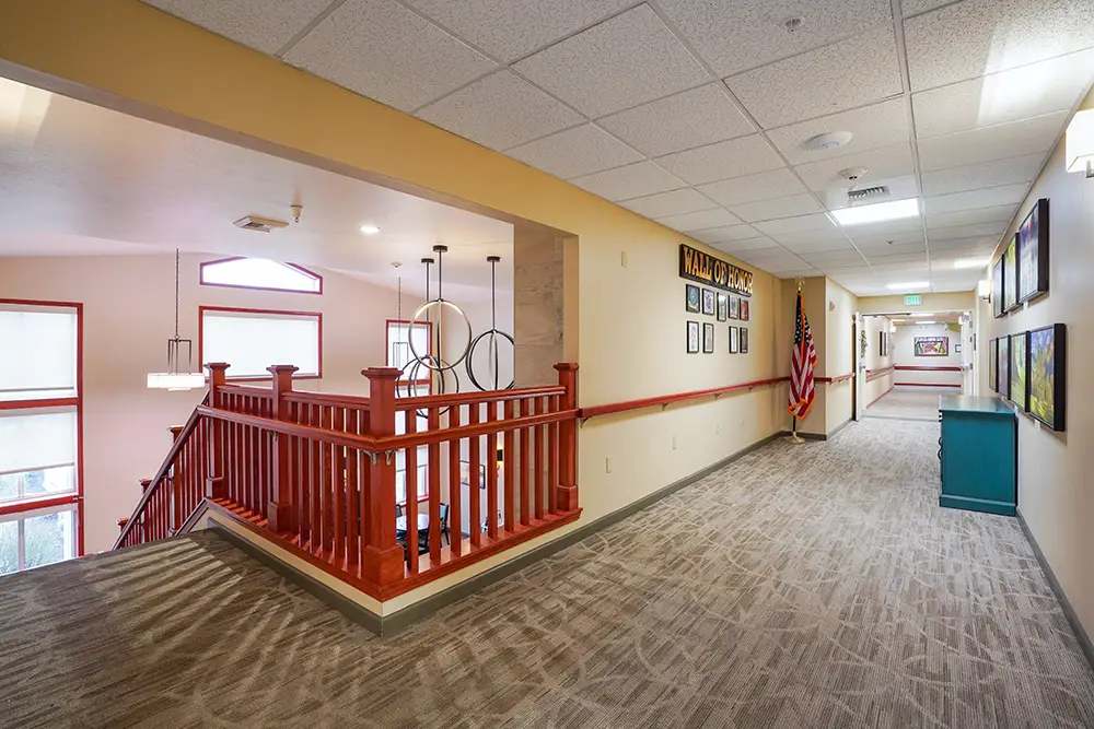 A carpeted hallway with framed art on the walls, a wooden stair railing on the left, and an American flag near a sign reading “Family Tree” at the far end. A carpeted hallway with framed art on the walls, a wooden stair railing on the left, and an American flag near a sign reading "Family Tree" at the far end.